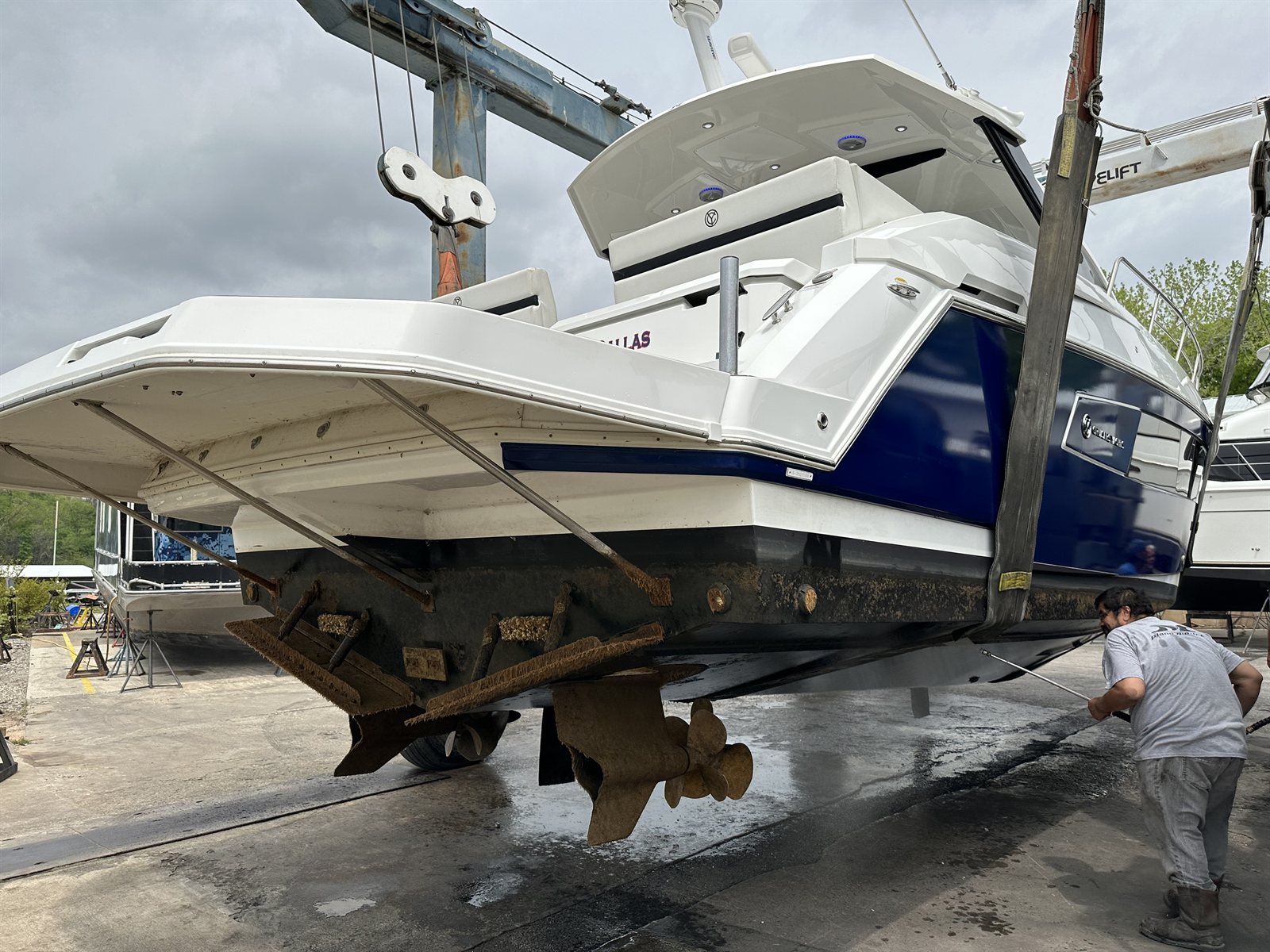 A cabin cruiser hauled out in travel-lift slings, its running gear heavily fouled with growth, being pressure-washed at a boatyard.